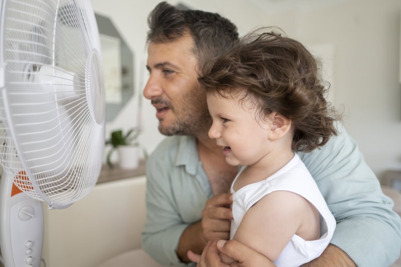father and toddler sitting in front of fan on a hot day.